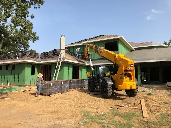 Construction workers installing shingles on a house roof, using a telehandler and ladder. Green siding visible.