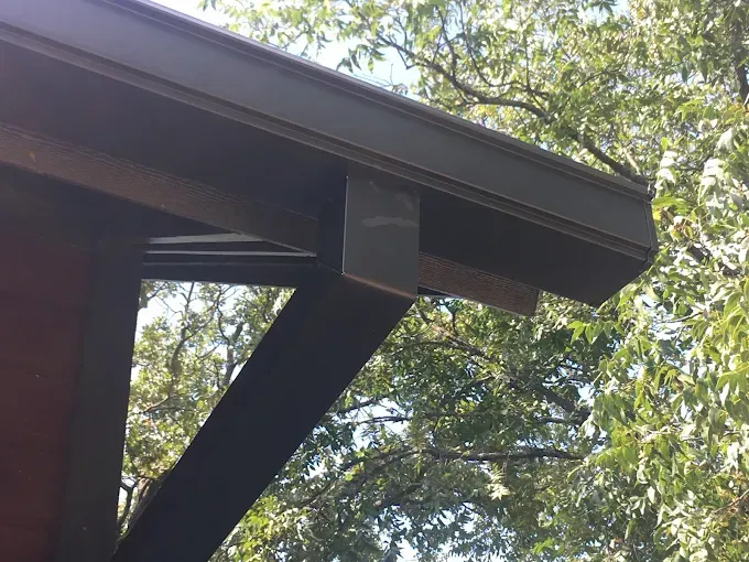 Dark brown rain gutter with a metal support bracket, attached to a brown wooden beam, surrounded by green trees.