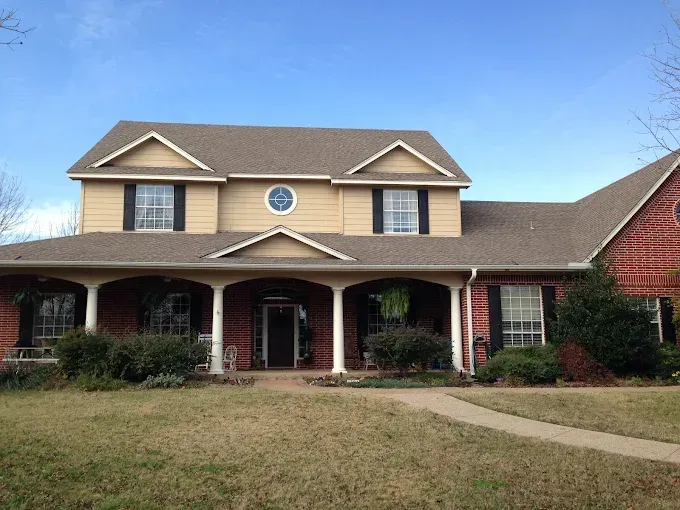 Two-story beige house with red brick accents, black shutters, and a covered porch on a sunny day.