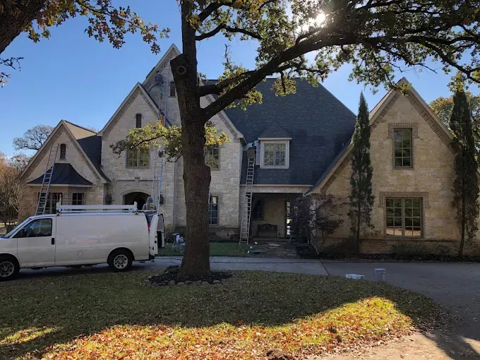 Large stone house with white van in the driveway; workers on ladders near the entrance and roof, under a tree.