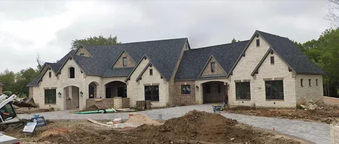 Large, light-colored stone house with dark roof under construction. Cloudy sky above dirt driveway.