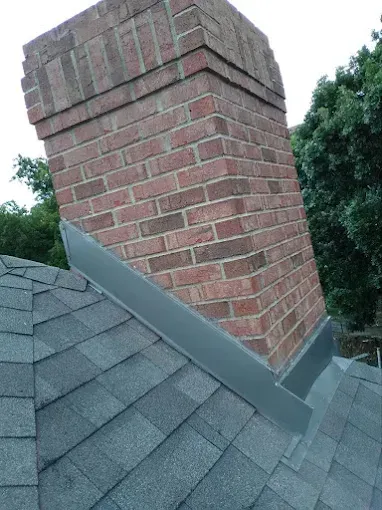 Brick chimney on a gray shingle roof, surrounded by flashing. Trees in the background.