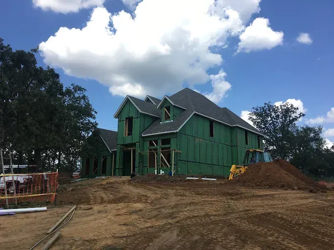 New house under construction on a dirt lot; green siding, dark roof, blue sky, and clouds.