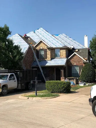 House with roof partially covered in blue underlayment; worker on the roof, ladder leans against it; truck nearby.