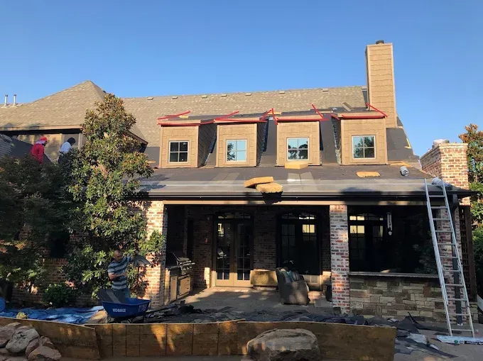 Rear view of a house under construction with brick, stone, and a chimney. Workers on the roof installing shingles.