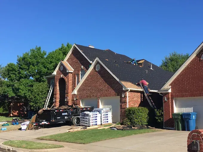 Roofers working on a brick house with a partially replaced roof; a dumpster and materials on the lawn.