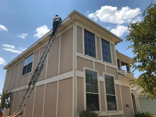 Man on a tall ladder cleaning gutters on a two-story beige house with white trim against a partly cloudy sky.