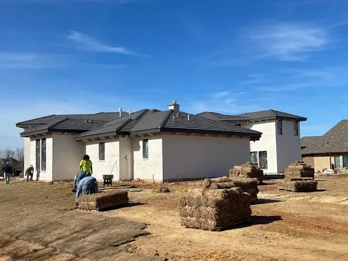 Workers installing sod around a stucco home with a dark roof under a blue sky.