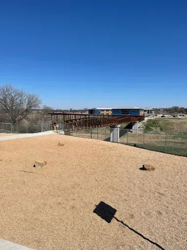 A sunny dog park with wood chip ground. Metal bridge in background. Fence, trees, and buildings visible.