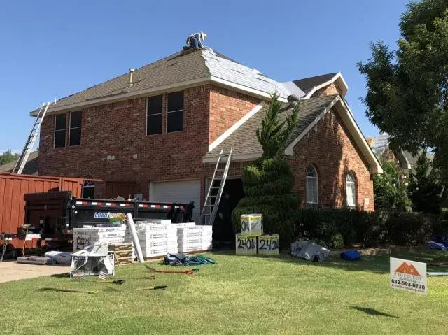 Roofing work on a two-story brick house. Workers on roof, materials and equipment in yard, ladders present.