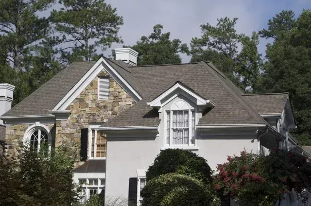 House with light-colored stone and stucco exterior, gray roof, and white trim. Trees in the background.