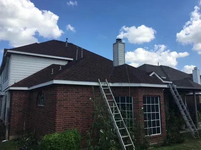 Brown shingle roof on brick house, with two ladders and chimney against a blue sky.