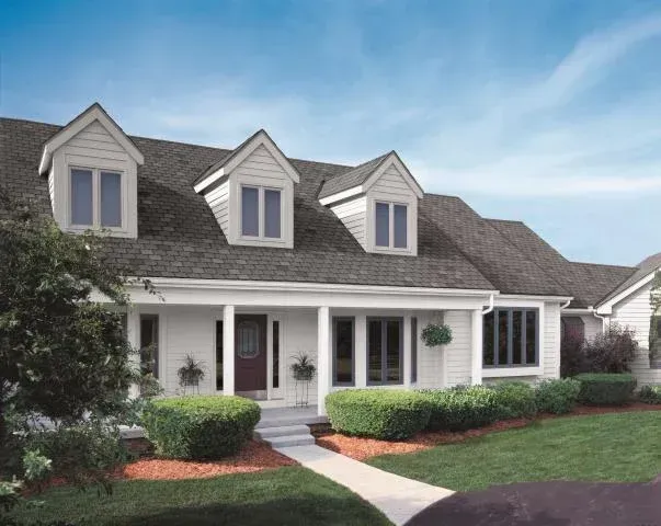 White house with a dark roof, three dormers, porch, bushes, and a green lawn. Blue sky.