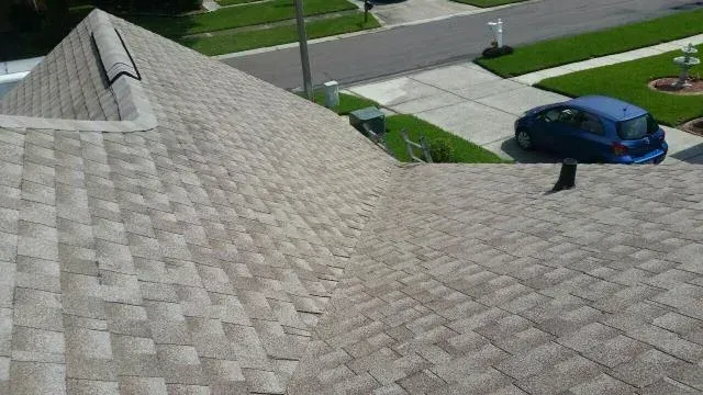 View from a roof with light-brown shingles. A driveway and blue car are in the background.