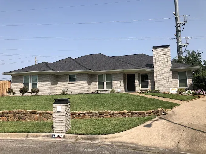 Gray brick ranch house with dark gray roof, chimney, and lush green lawn.