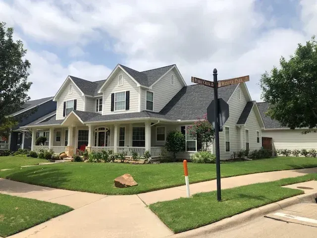 Two-story white house with black shutters, green lawn, sidewalk, and street sign on a sunny day.