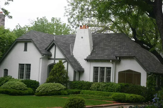 White Tudor-style house with dark gray roof, chimney, and landscaped front yard.