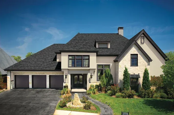 Two-story beige house with dark gray roof and garage doors, black trim, and front landscaping against a blue sky.