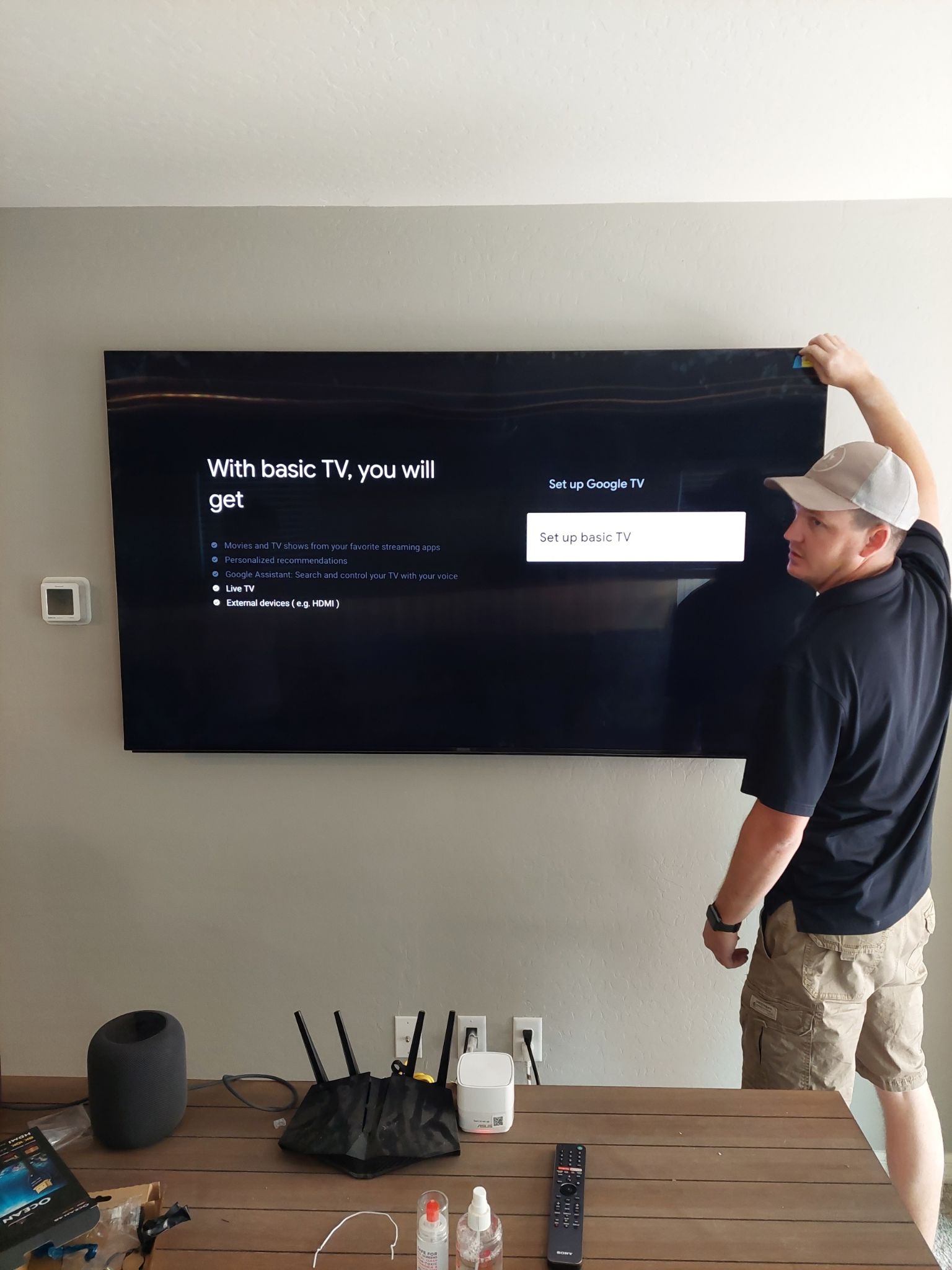 Man installing a TV on a wall; beige walls, dark TV, man in cap and shorts.