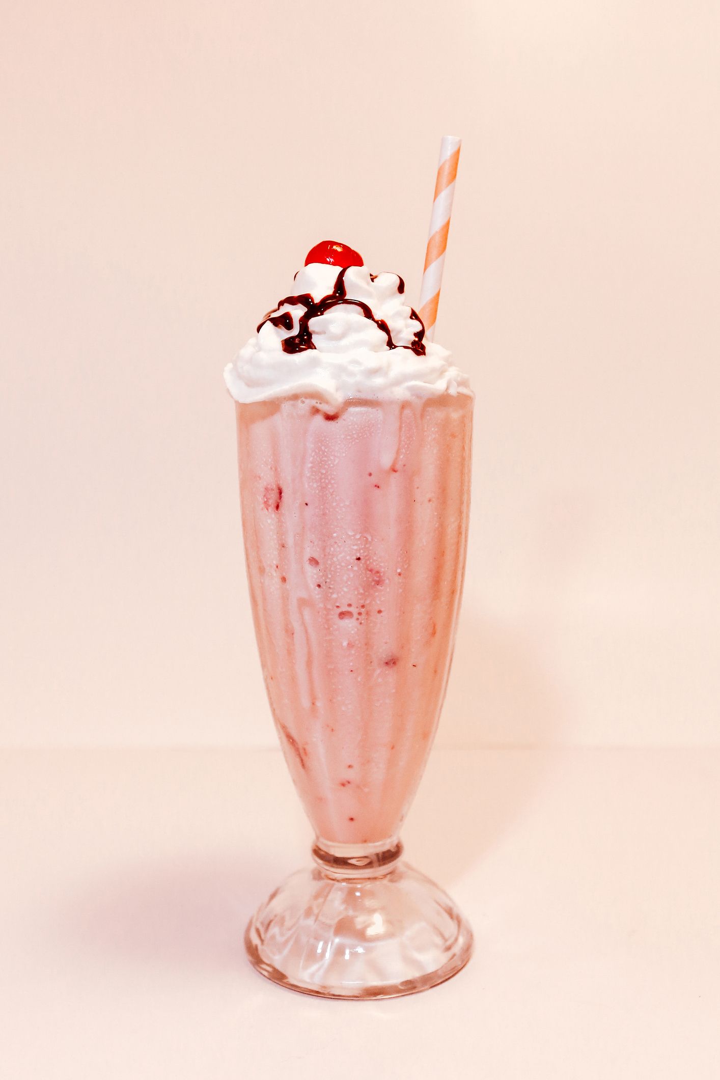 A strawberry milkshake in a tall glass, topped with whipped cream, chocolate drizzle, and a cherry, with a straw.