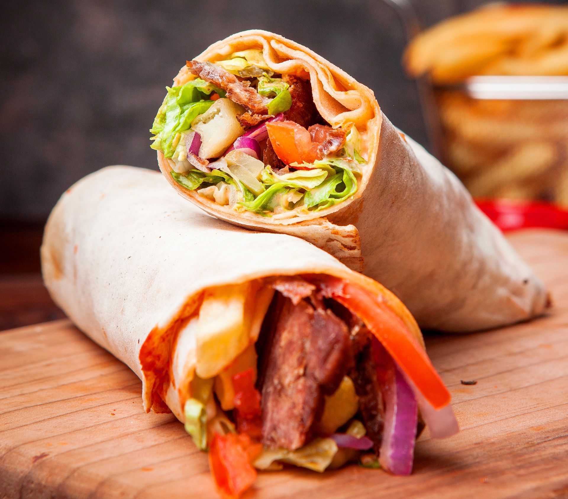 Two wraps filled with meat, fresh vegetables, and french fries sit on a wooden cutting board, with more fries in the back.