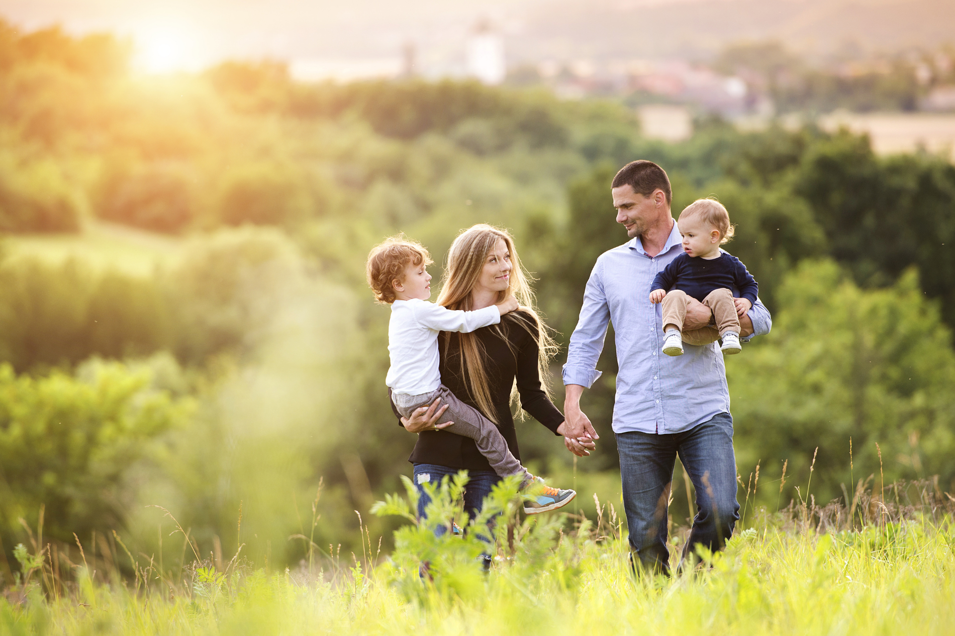 Family walking through a sunlit field with two children, holding hands and carrying a toddler