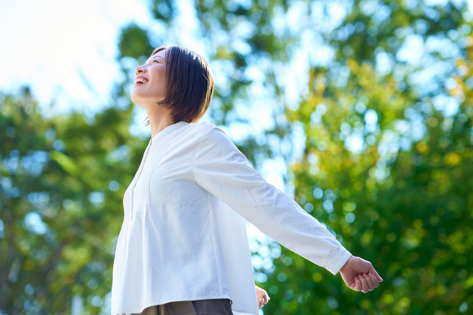 Person in a white shirt standing outdoors with arms outstretched, enjoying bright sunlight and greenery.