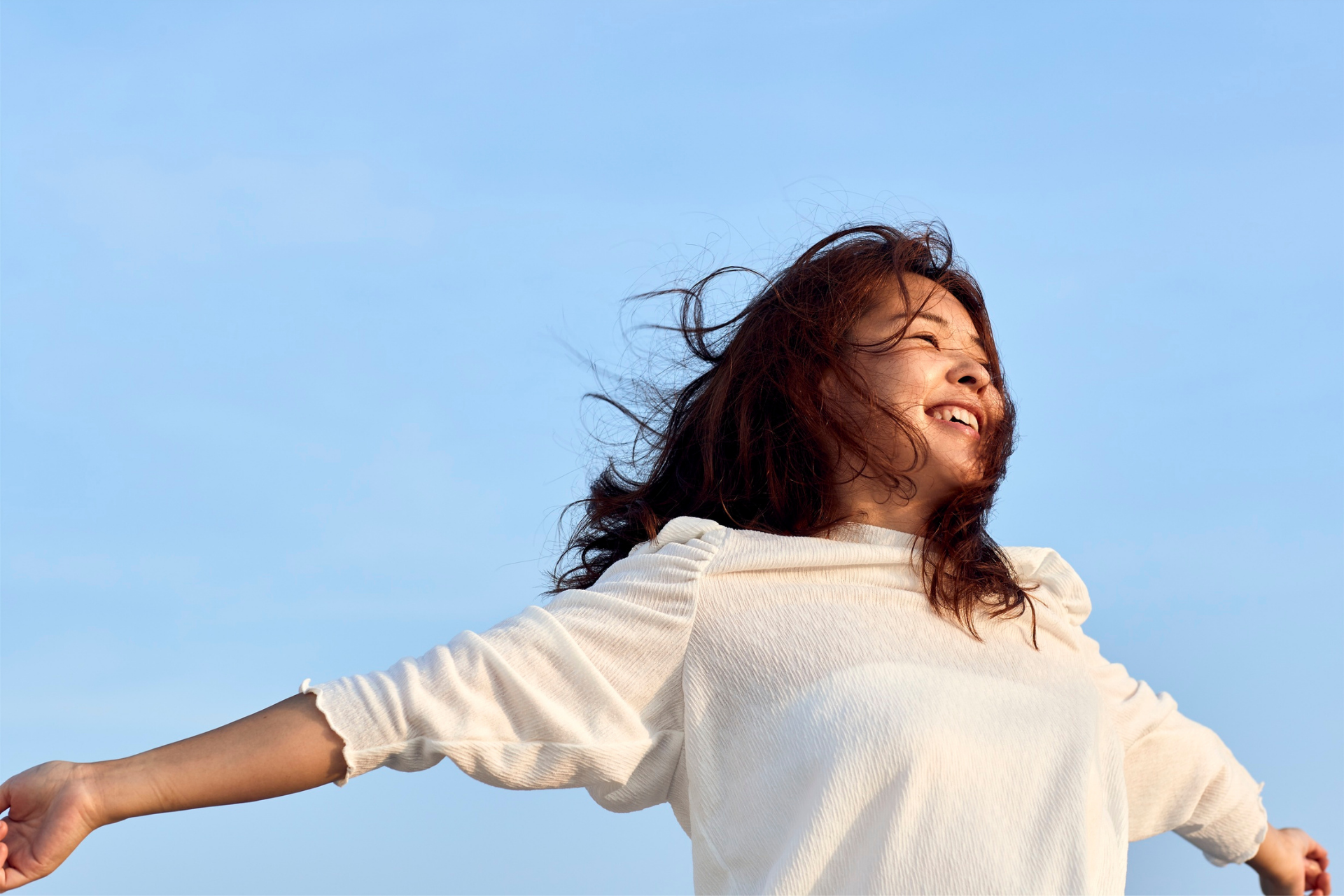 Person in a white shirt with arms outstretched, smiling against a blue sky.