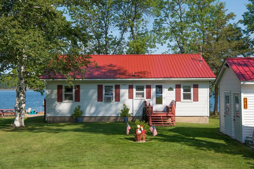 White cottage with a red roof by a lakeshore, with green lawn and trees in front