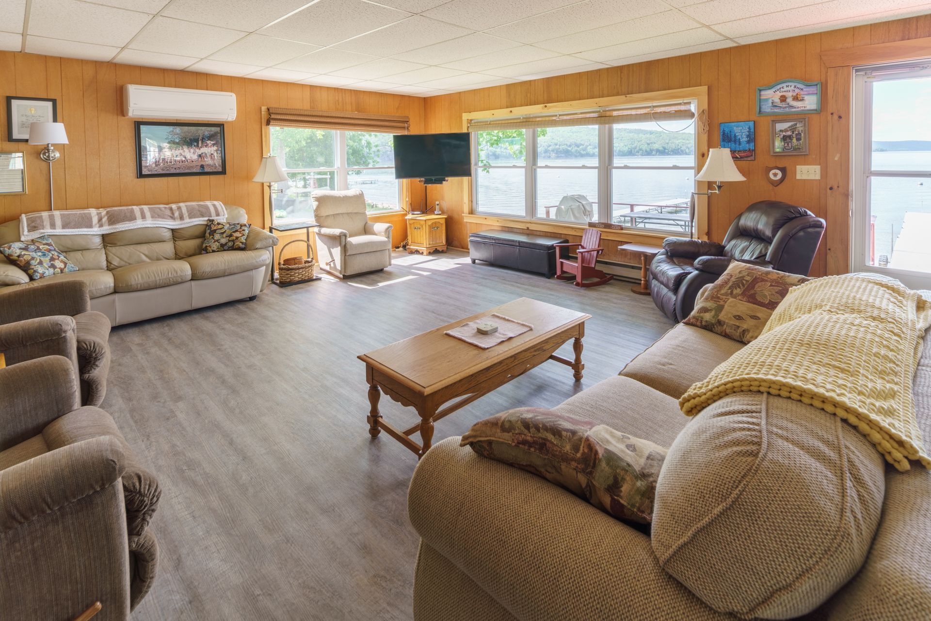 Spacious wood-paneled living room with beige sofas, coffee table, recliner, and large window overlooking water.