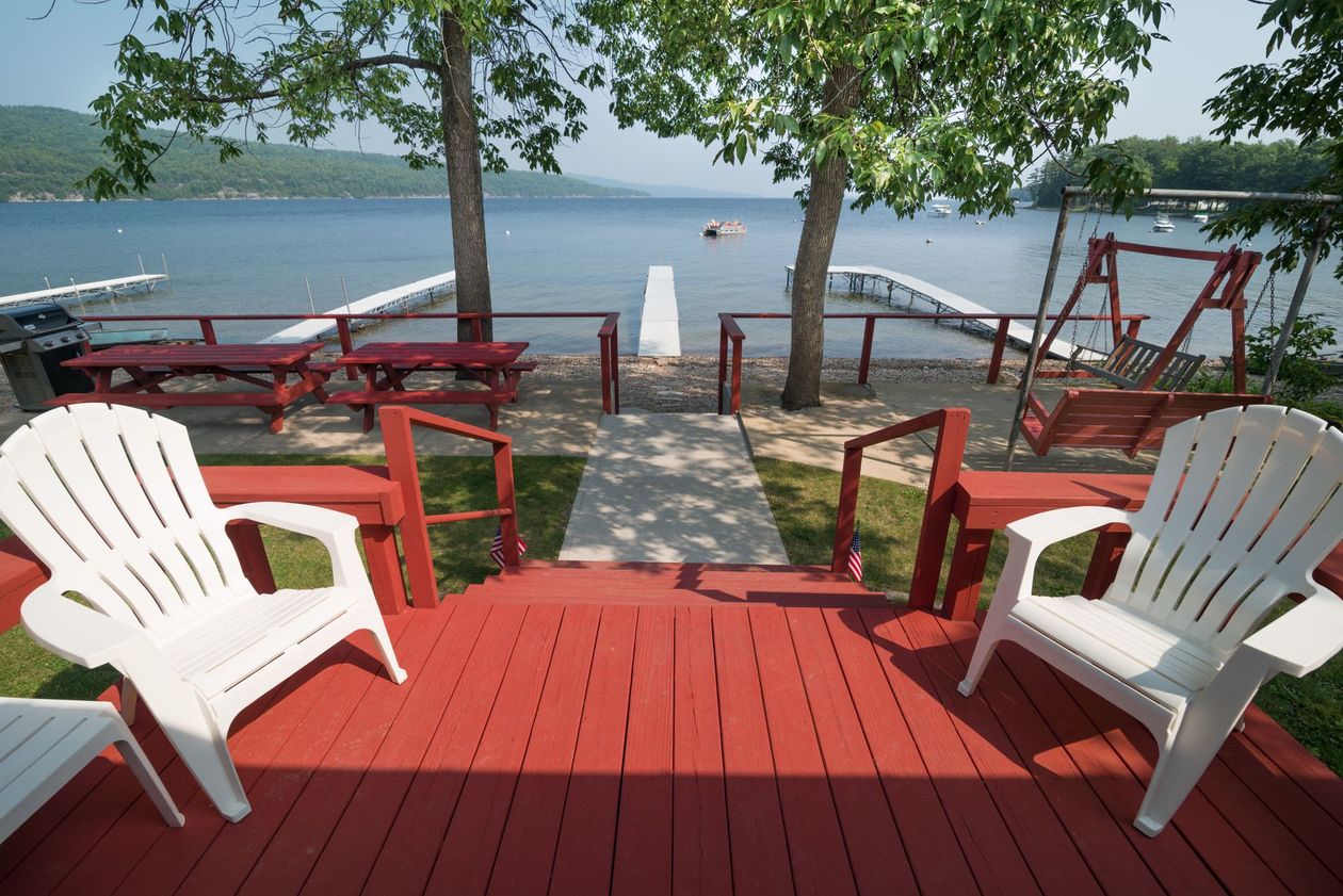 Red deck with white chairs overlooking a lake and dock under trees