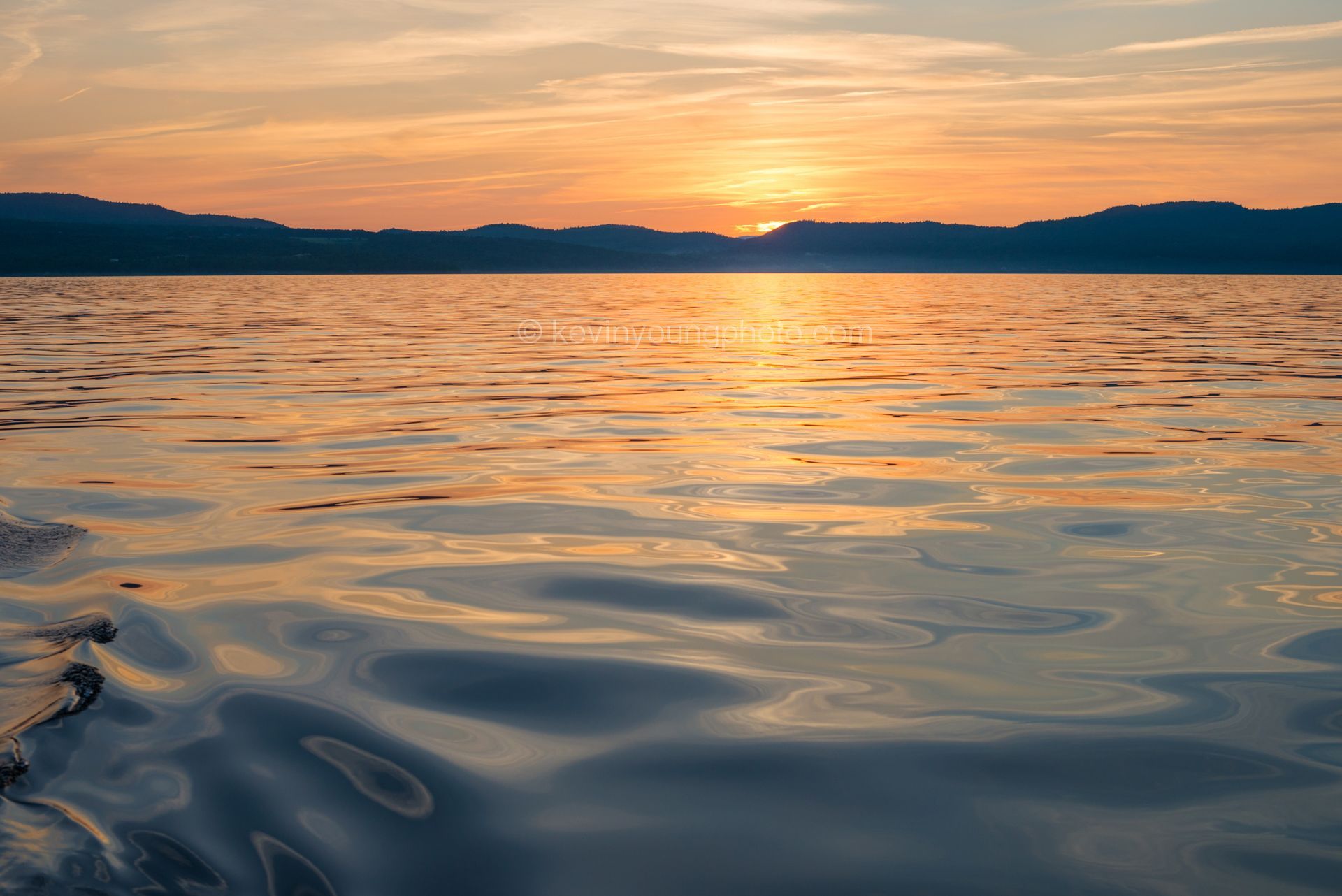 Sunset over a calm lake with orange light reflecting on rippling water and dark hills on the horizon