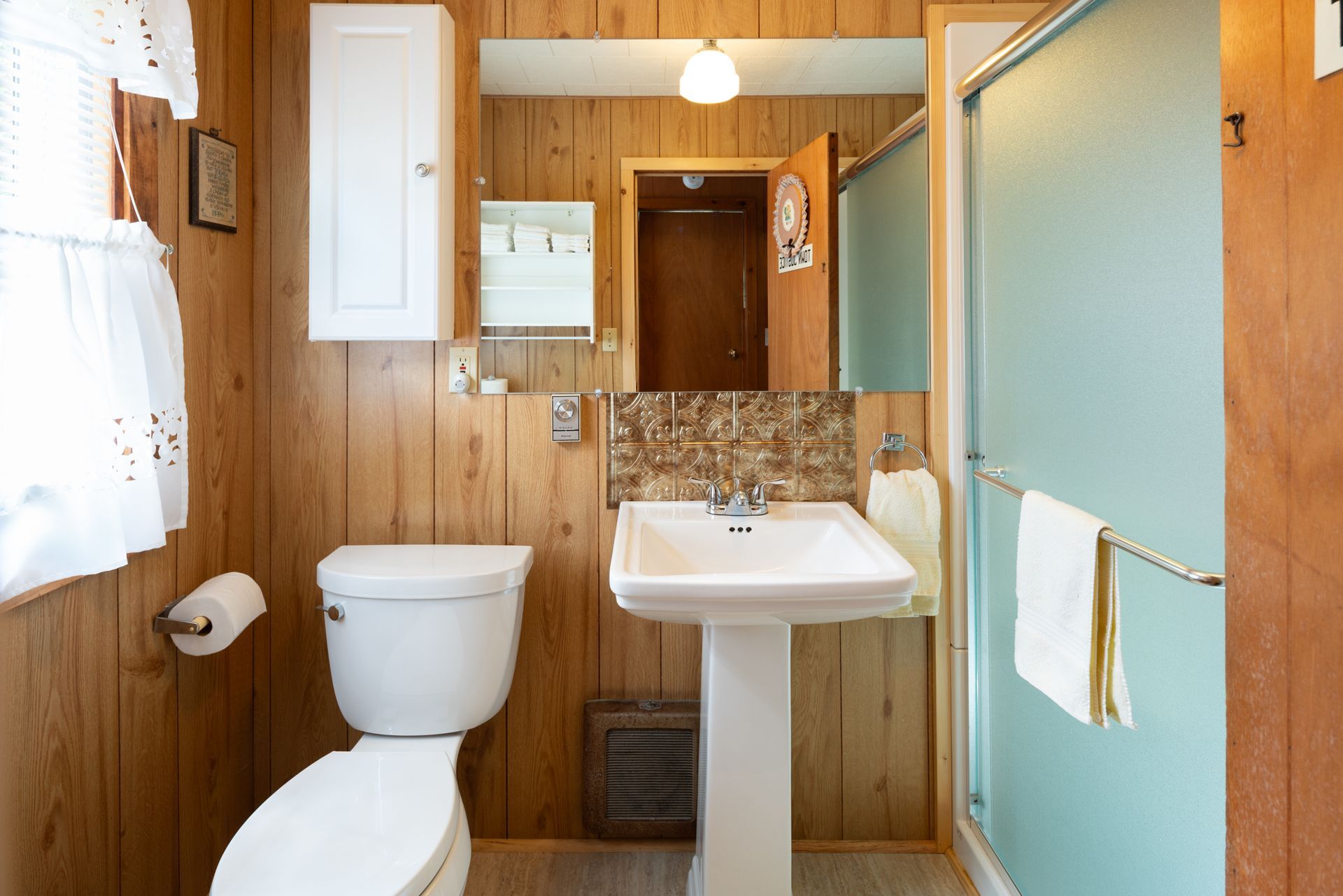 Small wood-paneled bathroom with toilet, sink, mirror, and frosted glass shower stall.