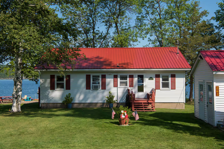 White cottage with red roof and shutters by a lake, with lawn, flowers, and trees nearby