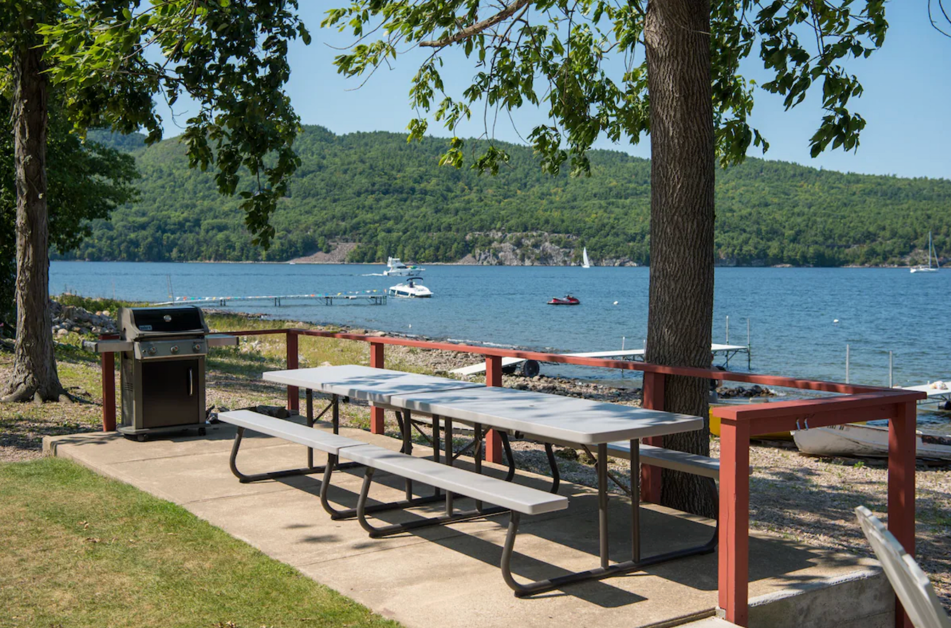 Picnic tables under trees by a lake with boats and wooded hills in the background