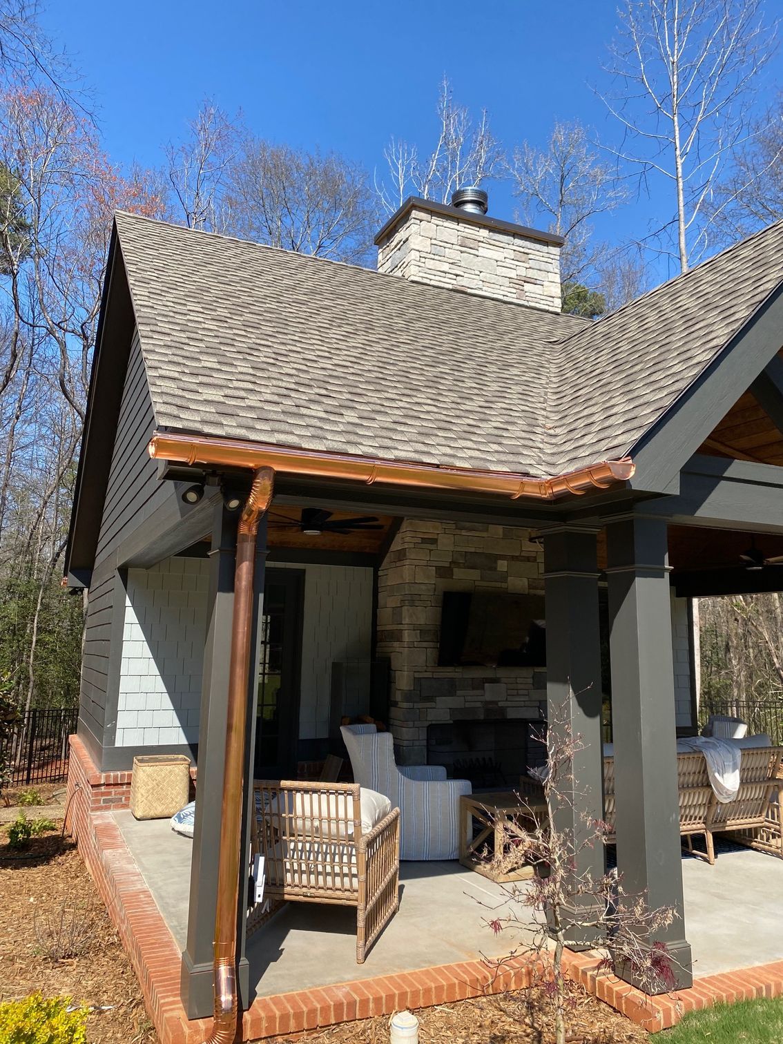 Stone porch with cushioned chairs under a shingled roof beside a brick-and-siding house on a sunny day