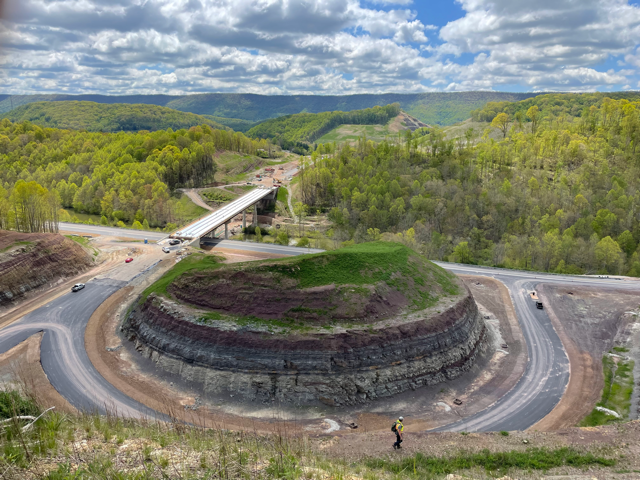 Highway construction project with forested hills in the background and a man in a safety vest in the foreground.