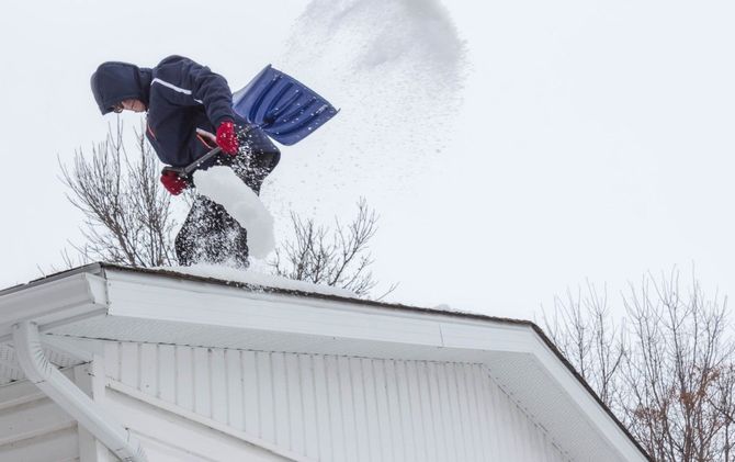Person shoveling snow off a white roof in winter, snow flying into the air