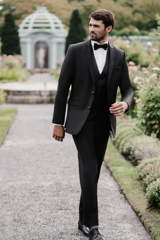 Man in a black tuxedo walking on a garden path with a white pavilion in the background
