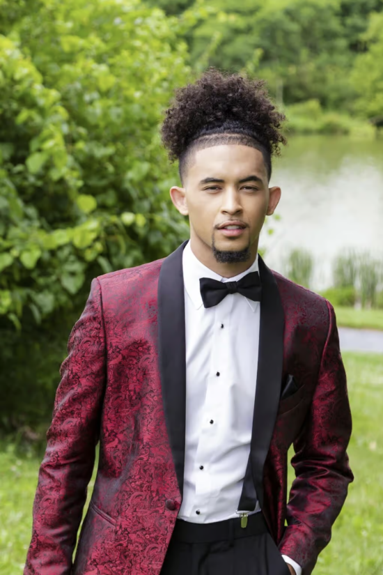 Man in a burgundy tuxedo jacket and black bow tie standing outdoors against green foliage