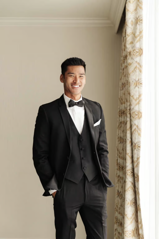 Man in a black tuxedo smiling by a window with patterned curtains