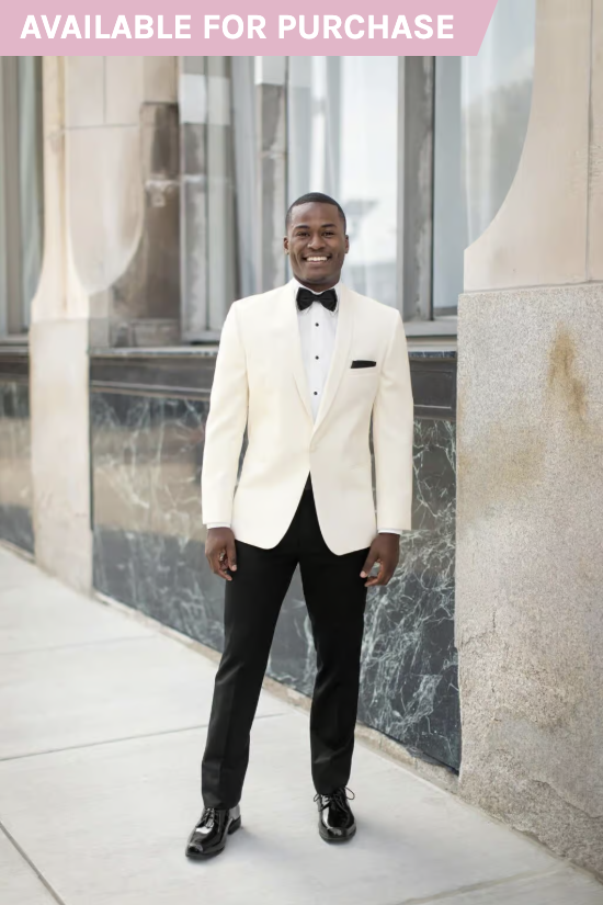 Man in a white tuxedo jacket and black trousers standing on a city sidewalk by a stone building.