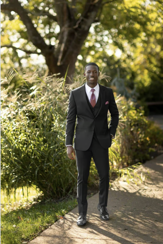 Man in a dark suit stands on a garden path with greenery and trees behind him.