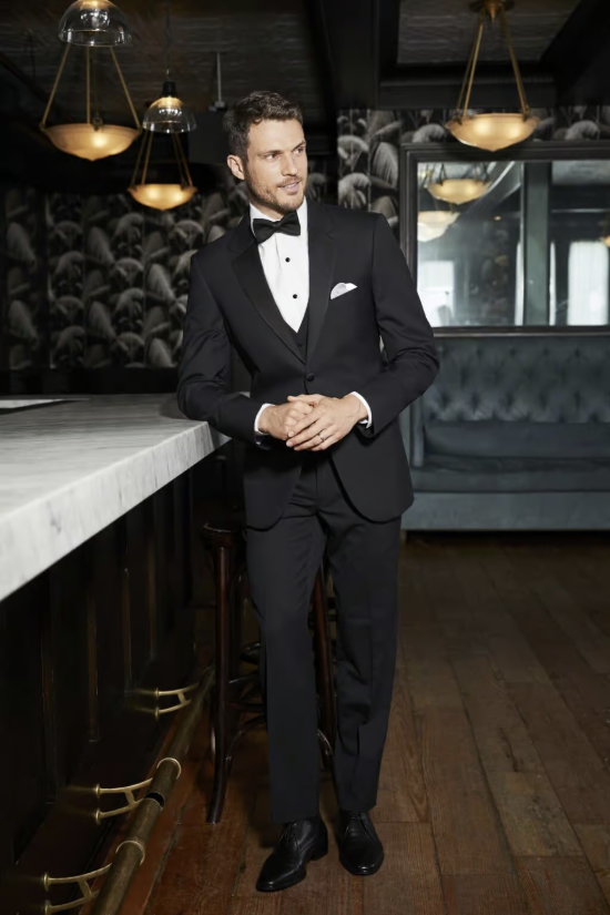 Man in a black tuxedo leaning against a bar in a dimly lit lounge