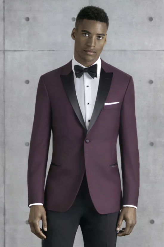 Man in a burgundy tuxedo jacket with black lapels and a bow tie standing against a concrete wall