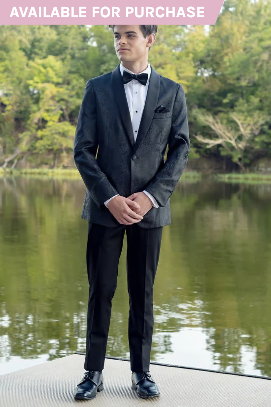 Man in a black tuxedo standing by a pond, with trees reflected in the water