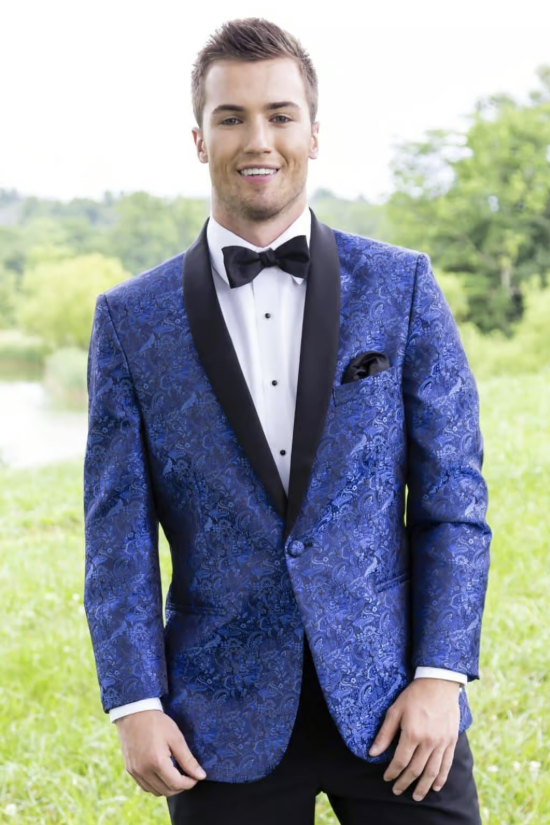 Man in a blue patterned tuxedo jacket and black bow tie standing outdoors, smiling.