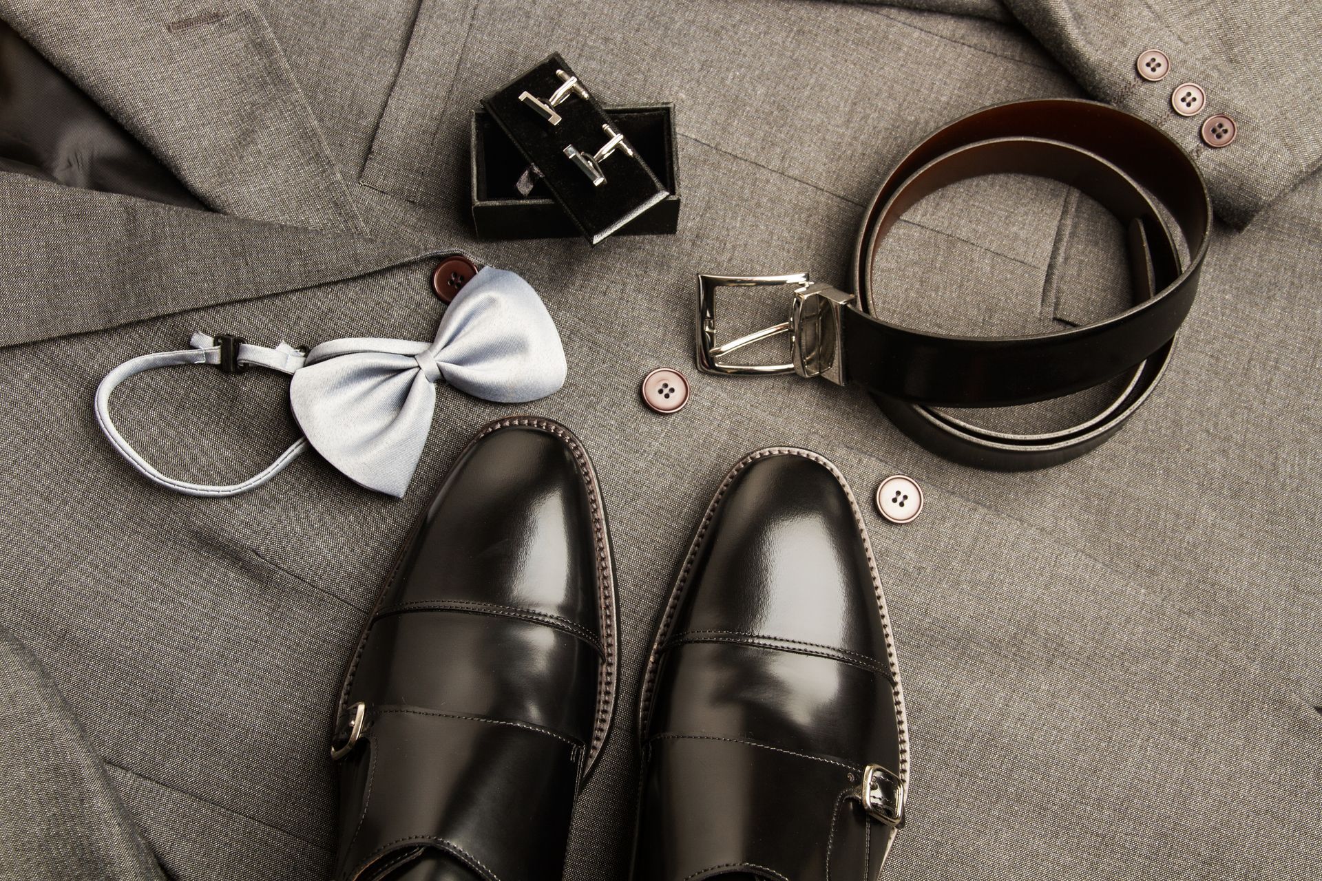 Men’s dress shoes, black bow tie, cufflinks, and belts on gray fabric.