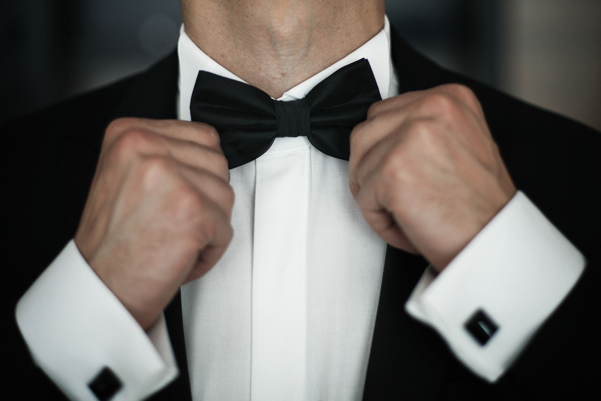 Man adjusting a black bow tie and tuxedo collar, wearing white shirt and cufflinks.