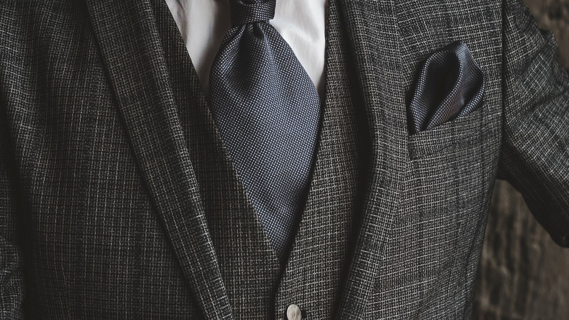 Close-up of a person wearing a dark textured suit jacket, gray tie, and pocket square.